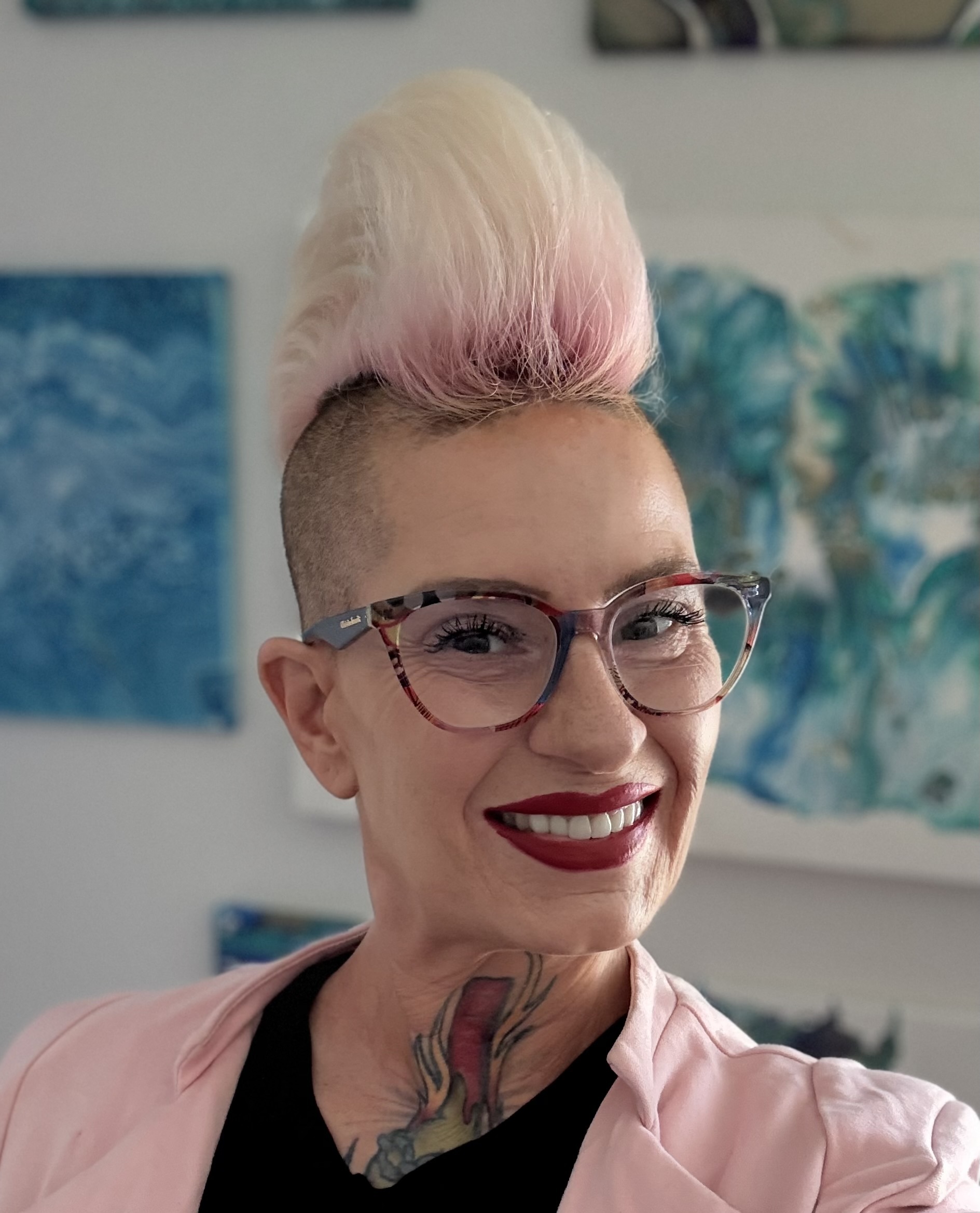 Picture os Shawna Stillwell with a pink pompadour hohawk, smiling warmly stanging in front of her paintings which she creates under the artist name Fluid Artist 805.